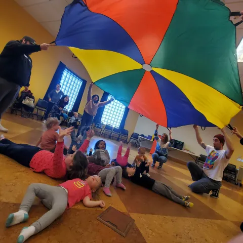 promotional image for Muncie Public Library's program "Tumbling Toddlers" held every Thursday at 11:30am at Kennedy Library, photo of adult library patrons holding a parachute up while children lay on the floor underneath the parachute